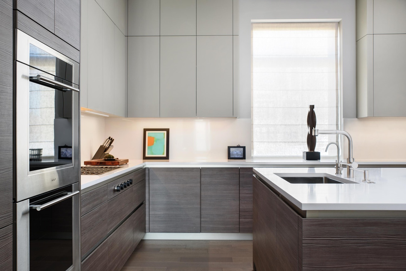 A modern kitchen with light gray upper cabinets, darker wood lower cabinets, white countertops, built-in oven, cooktop, and a window with natural light.